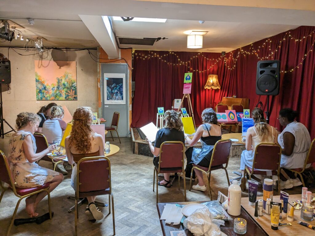 pairs of people painting canvases in from of a stage on which is placed various easels and pieces of artwork.