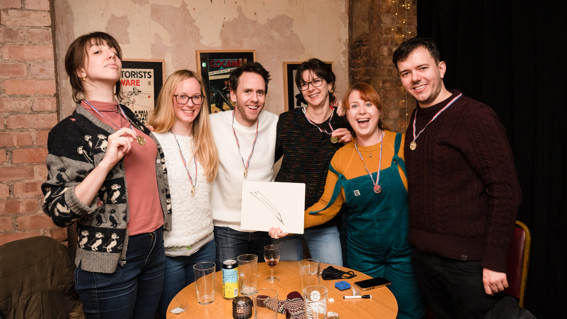 A group of six people at games night, standing around a table each wearing a medal