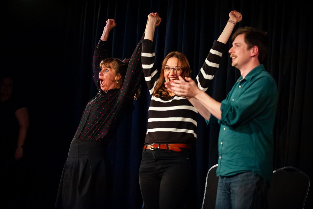 three members of the unscripted players. two women, one wearing a black long sleeve top and the other wearing a black and white striped jumper, holding hands with their arms in the air whilst a man in a green shirt claps next to them.