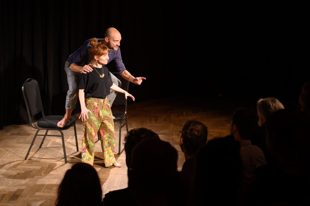 These Folk performing. Susan is stood barefoot on the floor, wearing a black t-shirt and colourful patterned trousers. Justin is stood on two chairs behind Susan and is grey trousers and a dark blue long sleeved t-shirt. 