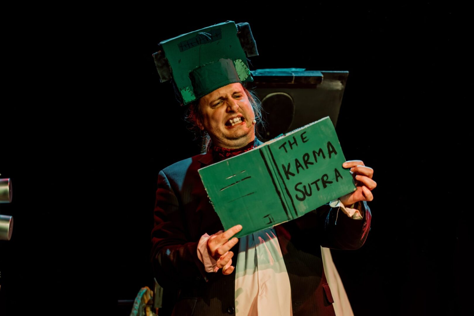 man with a green saucepan on his head reading a green book titled 'the karma sutra'