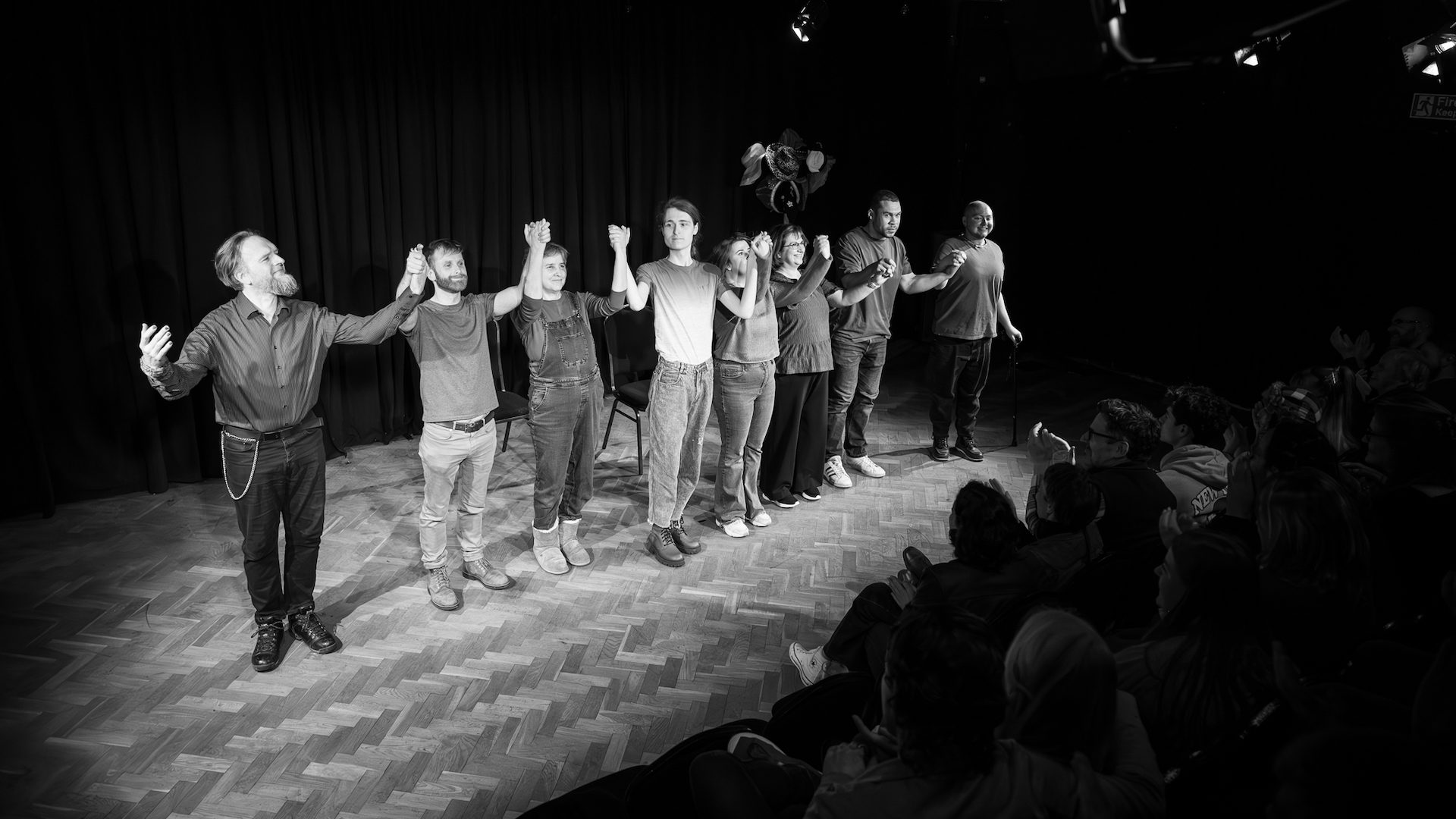 This is a black and white photo of a group of people stood on stage in a line, preparing to take a bow together.