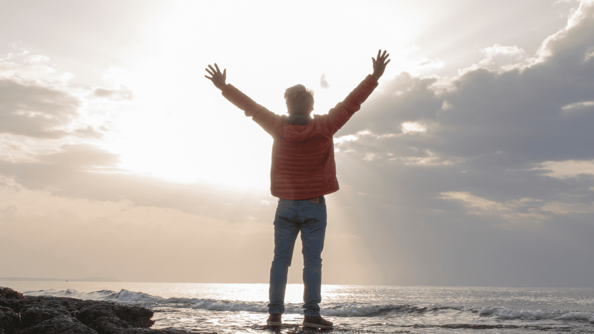 A man is seen from behind, standing on a beach with his hands raised to the sky