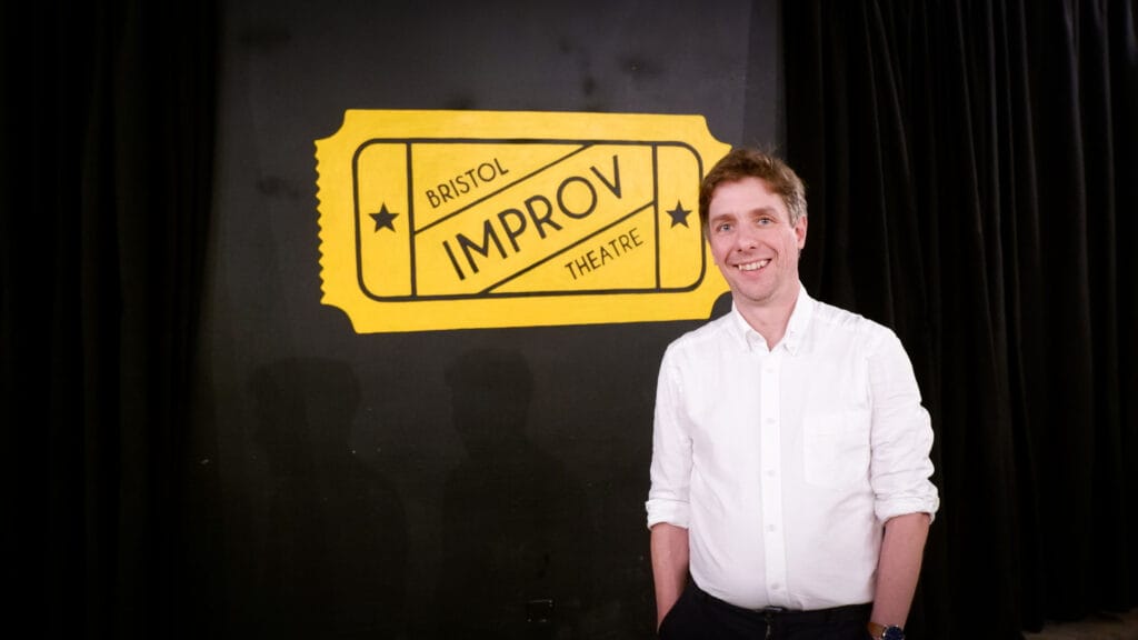 Bristol Improv Theatre's new CEO Tom Mansfield stands in front of a large painted yellow Bristol Improv Theatre logo. His hands are in his pockets and he smiles, looking at the camera.