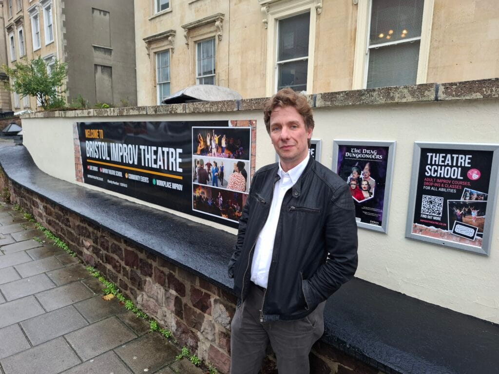 photo of man in jacket standing outside the sign for bristol improv theatre