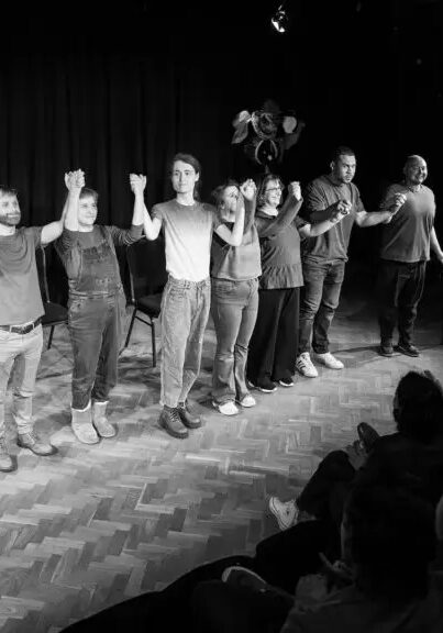 This is a black and white photo of a group of people stood on stage in a line, preparing to take a bow together.