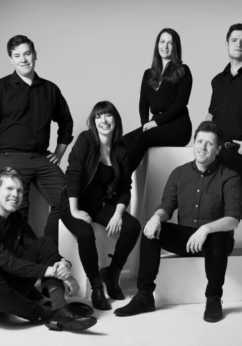 A black and white photo of 7 people stood and sat at different levels all smiling and looking at the camera.