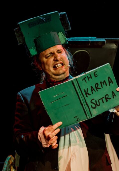 man with a green saucepan on his head reading a green book titled 'the karma sutra'