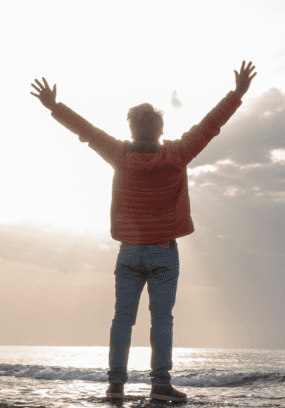 A man is seen from behind, standing on a beach with his hands raised to the sky